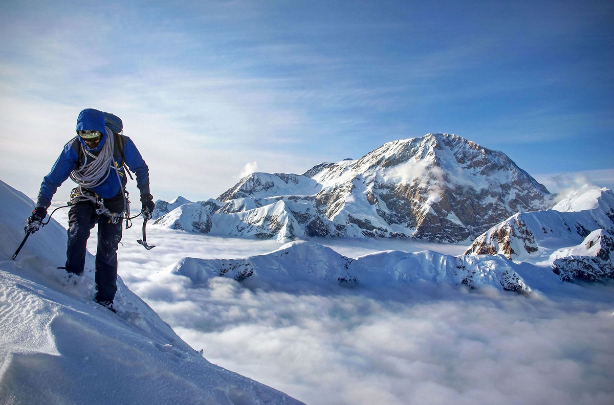 A mountaineer slowly makes his way across a snow field above the cloud line.