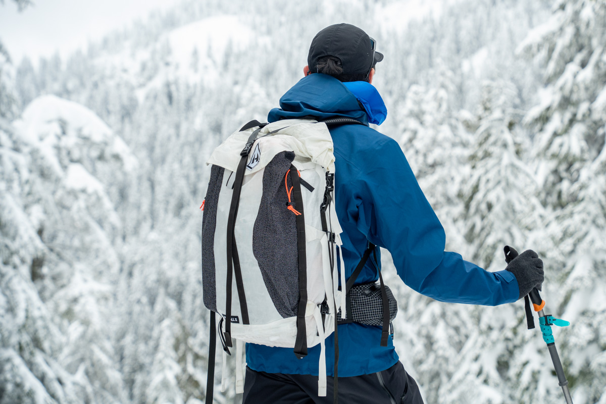 A man in a blue jacket faces a snowy forest landscape wearing a white ski backpack
