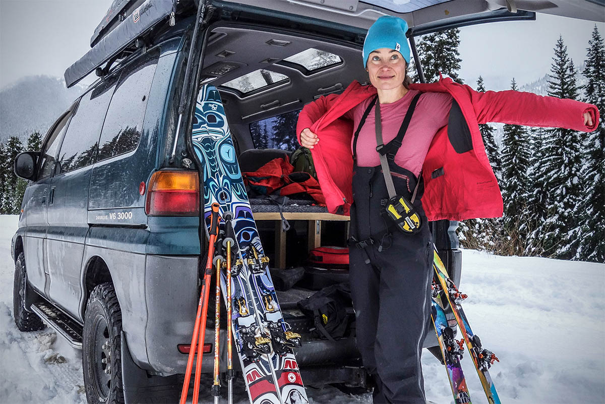 A women pulls on a jacket over her ski bibs and avalanche beacon that's strapped around her torso