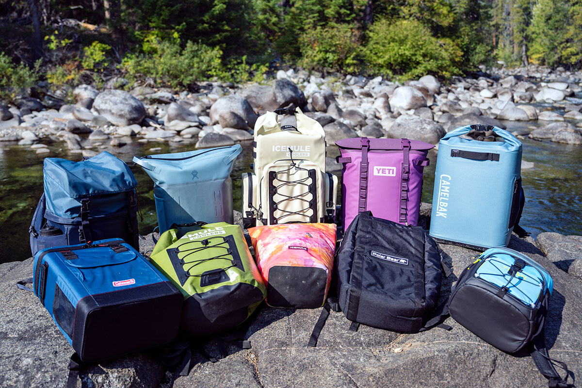 A display of ten backpack coolers in bright colors are lined up in a campground.