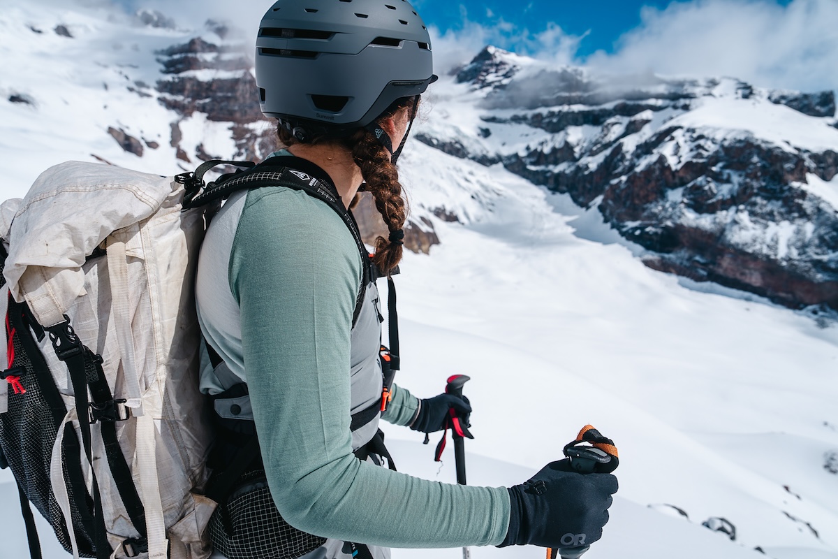 A woman stands in snowy mountains wearing a base layer, ski bibs, and a backpack