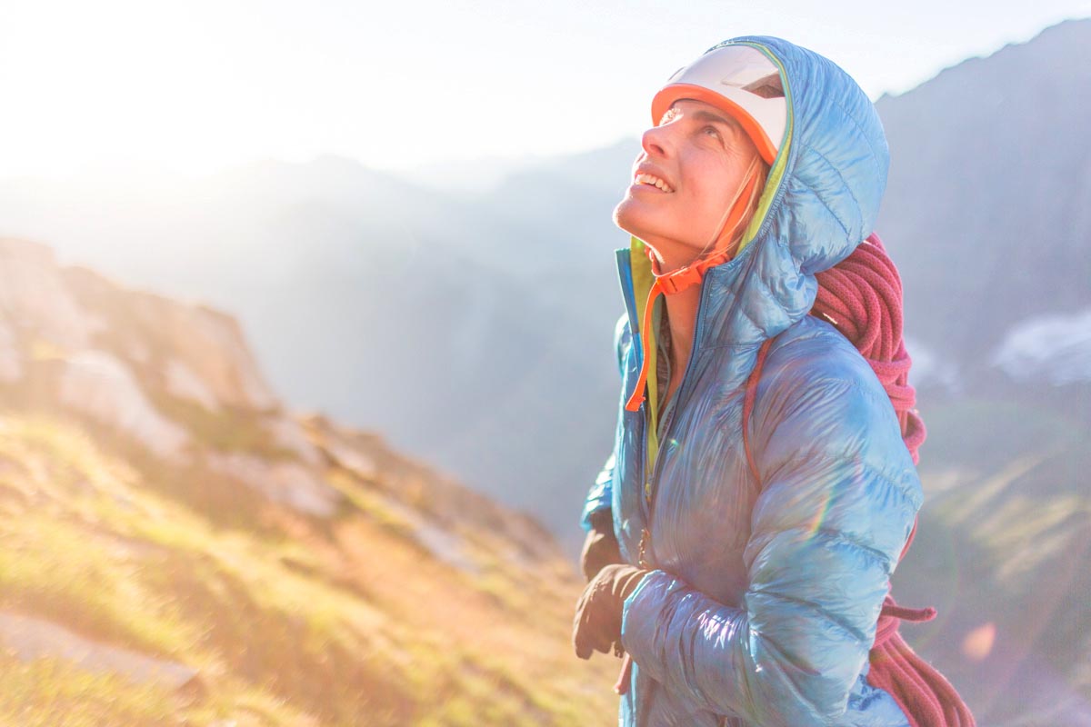 A woman wearing a climbing helmet and a hooded jacket looks up at a wall in the distance