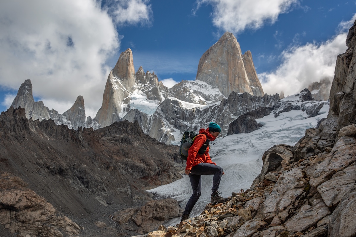 A woman hikes up steep rocks with jagged mountains and clouds in the background