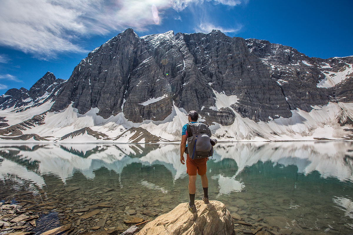 A man stands at the edge of a lake with large snowcapped mountains in the background