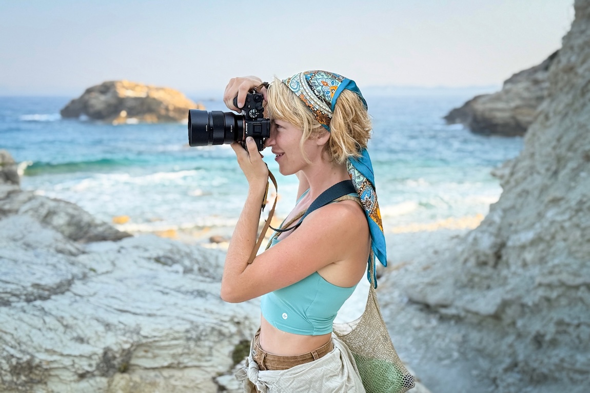 A woman stands next to the water looking through the lens of a camera