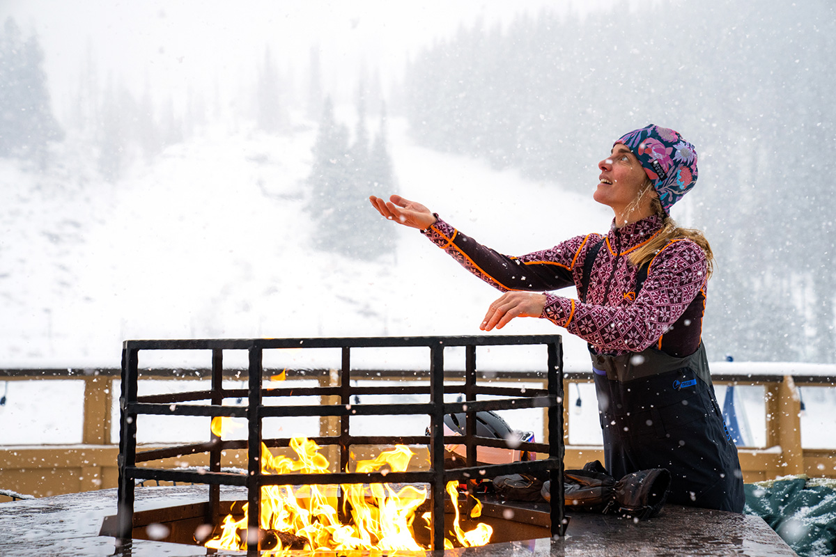 A woman standing by a fire on a snowy day wearing the Kari Traa Rose Half-Zip