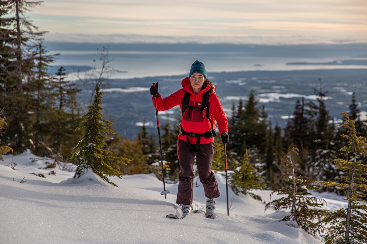 A woman skins up a snowy hill on skis wearing a red synthetic jacket