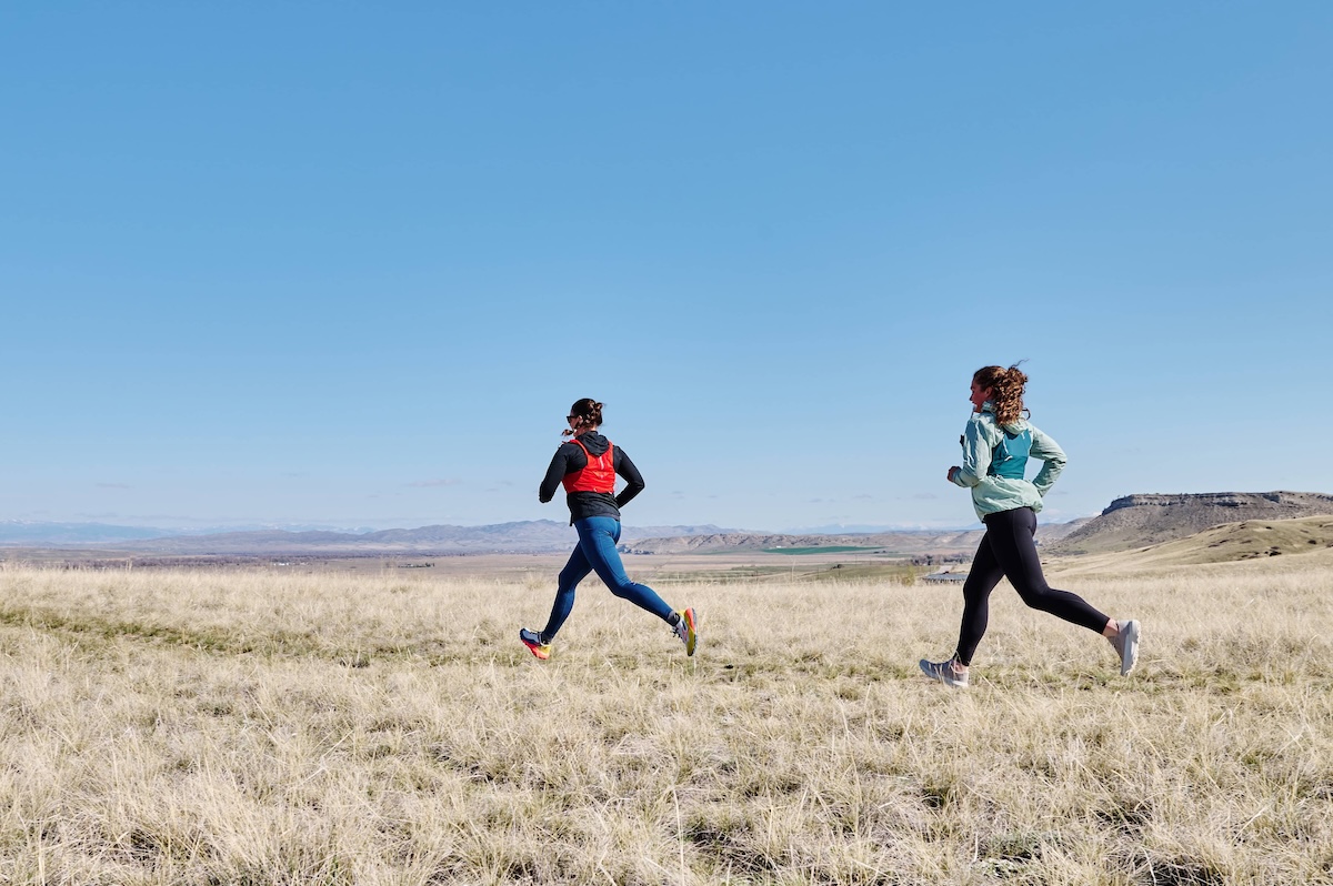 Two runners cross a field wearing running packs.