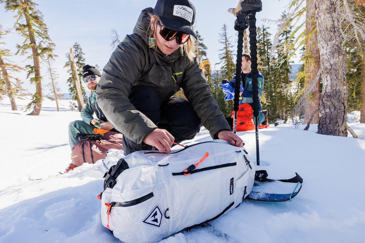 A skier kneeling down in the snow unzips a backpack