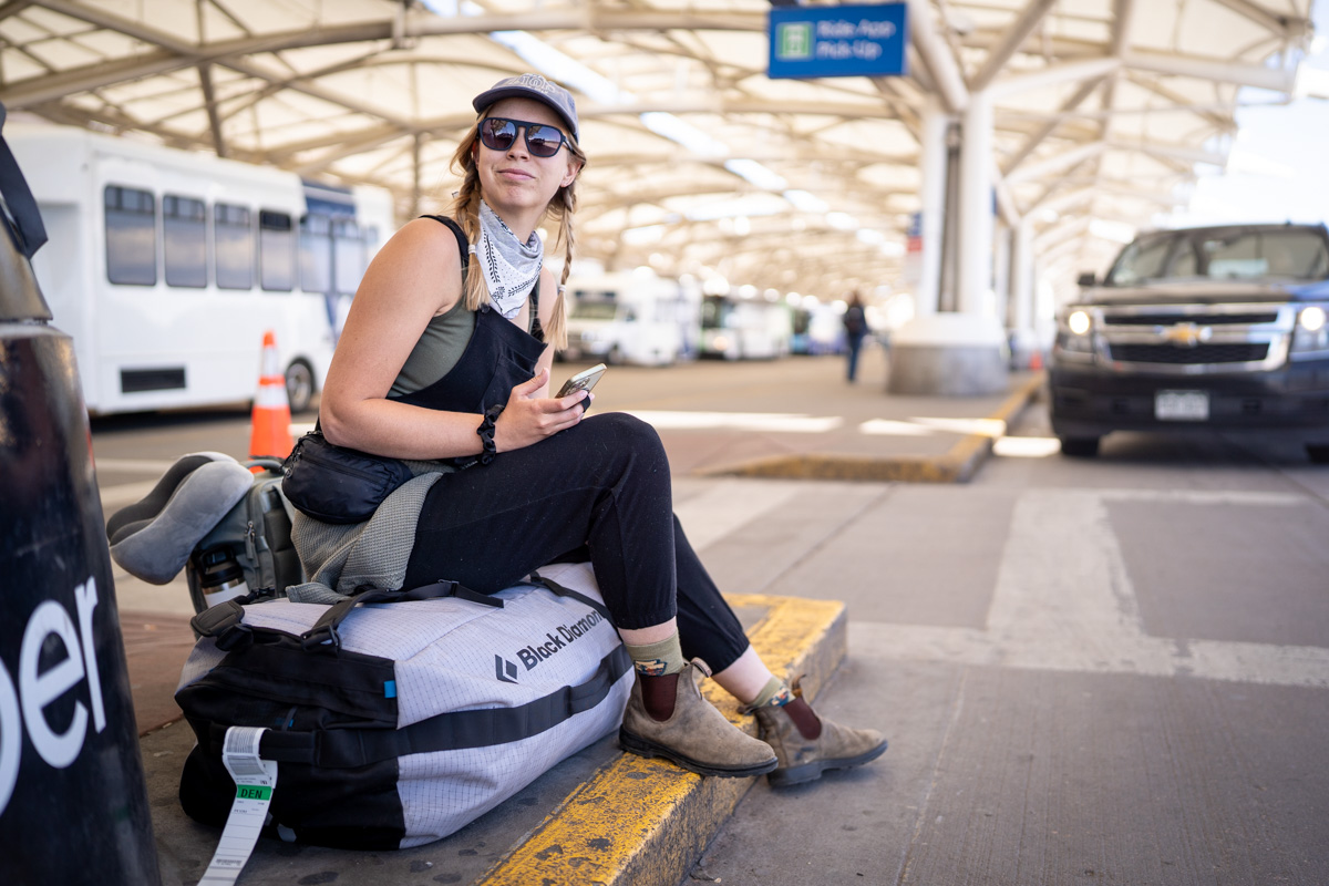 A woman sits on top of a duffel bag on the curb at the airport