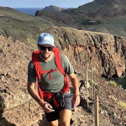 Matt walks up a steep hill with a backpack and the ocean in the distance