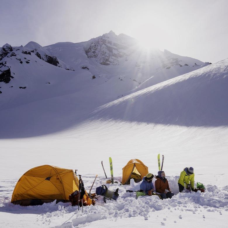 Three people sit in the snow next to tents and skis