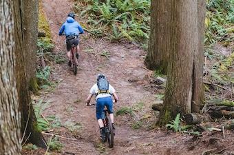 Two mountain bikers bike up a hill, one with a backpack and one with a waist pack