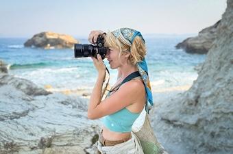 A woman stands next to the water looking through the lens of a camera