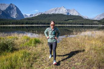 A woman in a fleece jacket walks toward us with a mountain range behind her