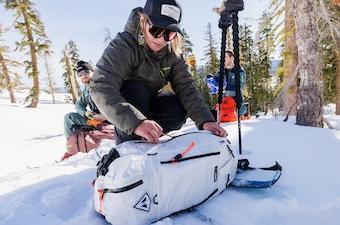 A skier kneeling down in the snow unzips a backpack